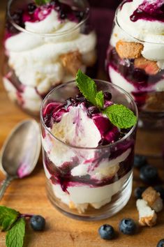 two glasses filled with ice cream and blueberries on top of a wooden table next to spoons