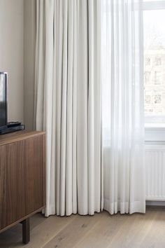 a television sitting on top of a wooden cabinet in front of a window with white curtains