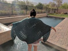 a woman is standing in the water with a shawl over her shoulders and looking at an outdoor swimming pool