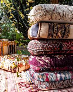 a pile of pillows sitting on top of a colorful rug next to a tree and bushes