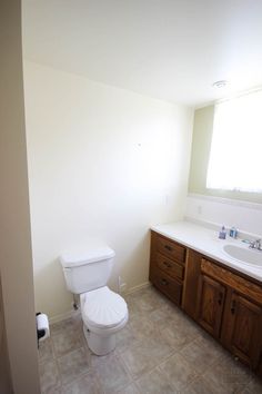 a white toilet sitting next to a sink in a bathroom under a window on top of a tiled floor