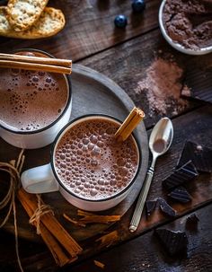 two cups of hot chocolate on a plate with cinnamon sticks and blueberries next to them