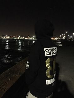 a person wearing a black hoodie standing on a pier at night with the city lights in the background