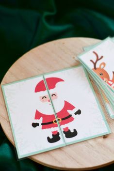 two christmas cards with santa claus on them sitting on top of a wooden table next to a green cloth