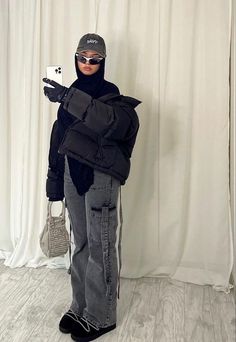 a person in black jacket and hat holding up a cell phone while standing next to a white curtain