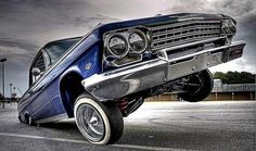 an old blue car is parked in a parking lot with cloudy skies behind it and the front lights are on