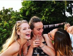 three young women are laughing and drinking wine at an outdoor party with bunting streamers in the background