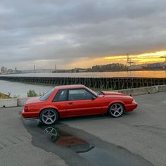 a red car parked in front of a body of water with the sun setting behind it