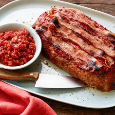 a white plate topped with meat covered in sauce next to a bowl of salsa and a knife