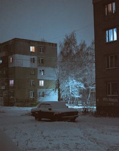 an old car is parked in the snow by some buildings at night time with lights on