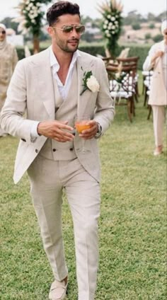 a man in a suit and sunglasses is walking through the grass with an orange drink