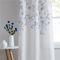 a white vase with blue flowers sitting next to a curtain in front of a window