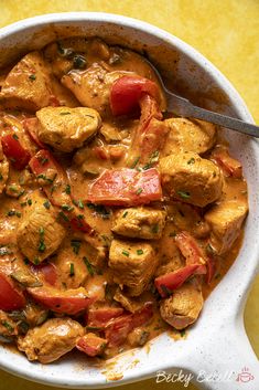 a white bowl filled with chicken and tomatoes on top of a yellow table cloth next to a spoon