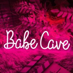 a pink neon sign that says bake cave on top of a furry rug next to flowers