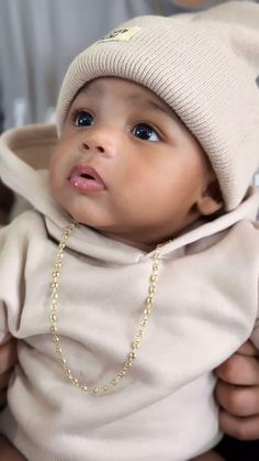 a baby wearing a sweater and a beanie looks up at the camera while being held by its mother