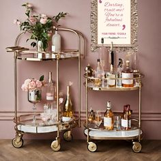 two gold drinks trolleys with bottles and glasses on them in front of a pink wall