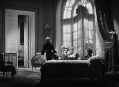 an old fashioned bedroom with two women sitting at the foot of a bed in front of a large window