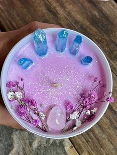 a hand holding a bowl filled with blue and pink liquid next to purple flowers on top of a wooden table