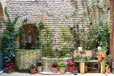 an outdoor fountain surrounded by potted plants in front of a brick wall and doorway