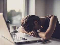 a woman leaning her head on the desk next to a laptop computer and coffee cup