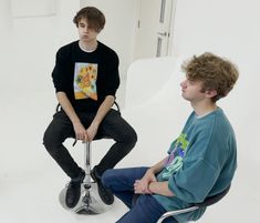 two young men sitting on top of chairs in front of a white wall and one is talking to the other