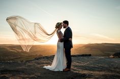 a bride and groom standing on top of a hill at sunset with their veil blowing in the wind