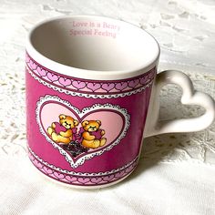 a pink coffee mug with two teddy bears in the center on a white lace tablecloth