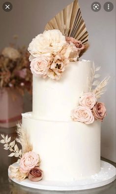 a three tiered white cake with pink flowers and feathers on top is sitting on a table