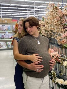 a pregnant woman hugging her husband in a flower shop with flowers on the shelves behind her