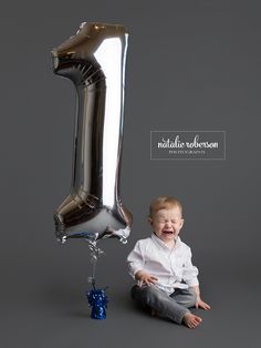 a baby sitting on the ground next to a large silver number one balloon that says 1