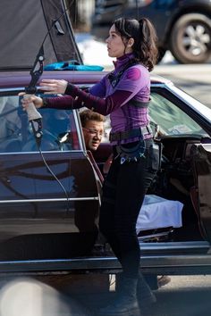 a woman standing next to a car with her hand on the door and another person sitting in the driver's seat