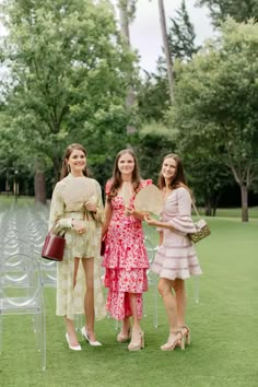 three women standing next to each other on a grass covered field with chairs and trees in the background