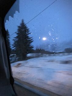 the view from inside a vehicle looking out at snow covered ground and pine trees in the distance