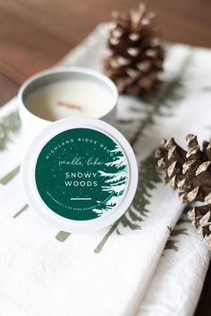 a candle and some pine cones on a table with a white towel that says snowy woods