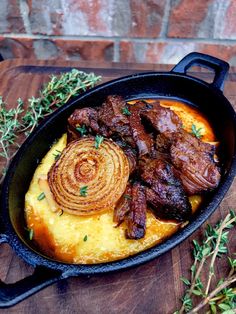 a skillet filled with meat and potatoes on top of a wooden table next to herbs