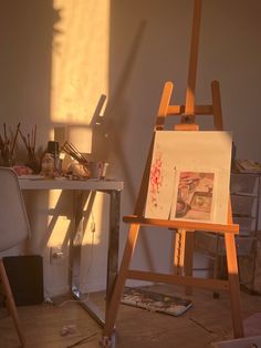 an easel is sitting next to a desk with art supplies on it and the sun shining through the window