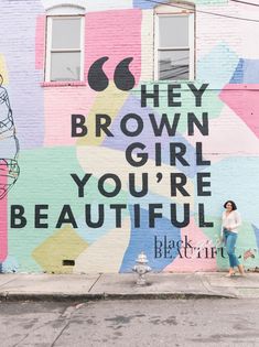 a woman standing in front of a colorful wall with the words hey brown girl you're beautiful painted on it