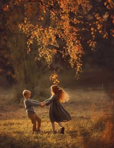 two young children holding hands under a tree in the fall time with leaves on it