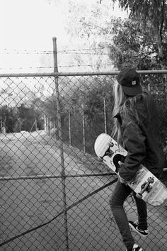 a person holding a skateboard in front of a chain link fence with trees behind them