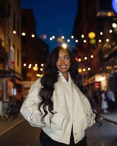 a woman standing in the middle of a street at night wearing a white jacket and black pants