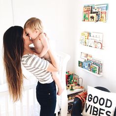 a woman holding a baby in her arms and kissing the child's cheek while standing next to a white crib