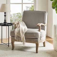 a chair with a blanket on it next to a lamp and table in front of a window