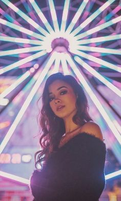 a woman standing in front of a ferris wheel at night with bright lights behind her