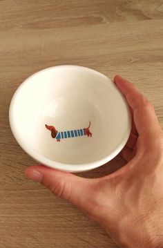 a hand holding a white bowl with a red and blue dachshund on it