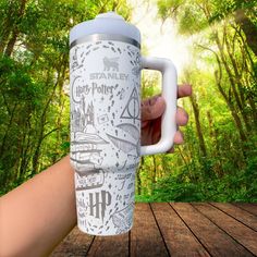 a hand holding a white travel mug with harry potter symbols on it in the woods