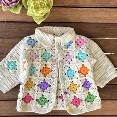 a white crocheted jacket with colorful flowers on the front and bottom, sitting on a wooden surface