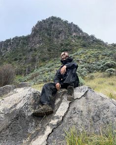 a man sitting on top of a large rock in the middle of a field next to a mountain