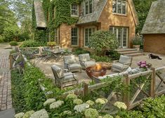 a patio with chairs and fire pit in front of a brick house surrounded by greenery