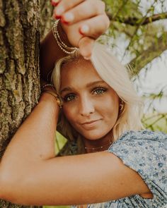 a woman leaning against a tree with her hands on her head and looking at the camera