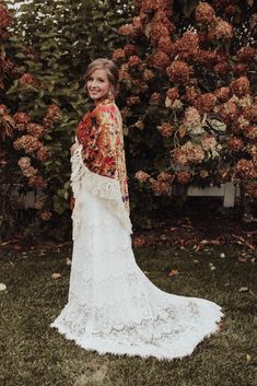 a woman standing in front of a bush wearing a white lace dress and red shawl